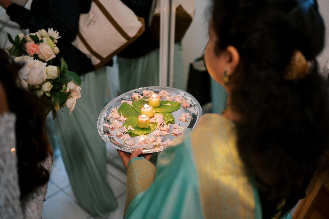 Woman holding a decorative tray with lit candles, flower petals, and green leaves during a traditional ceremony.