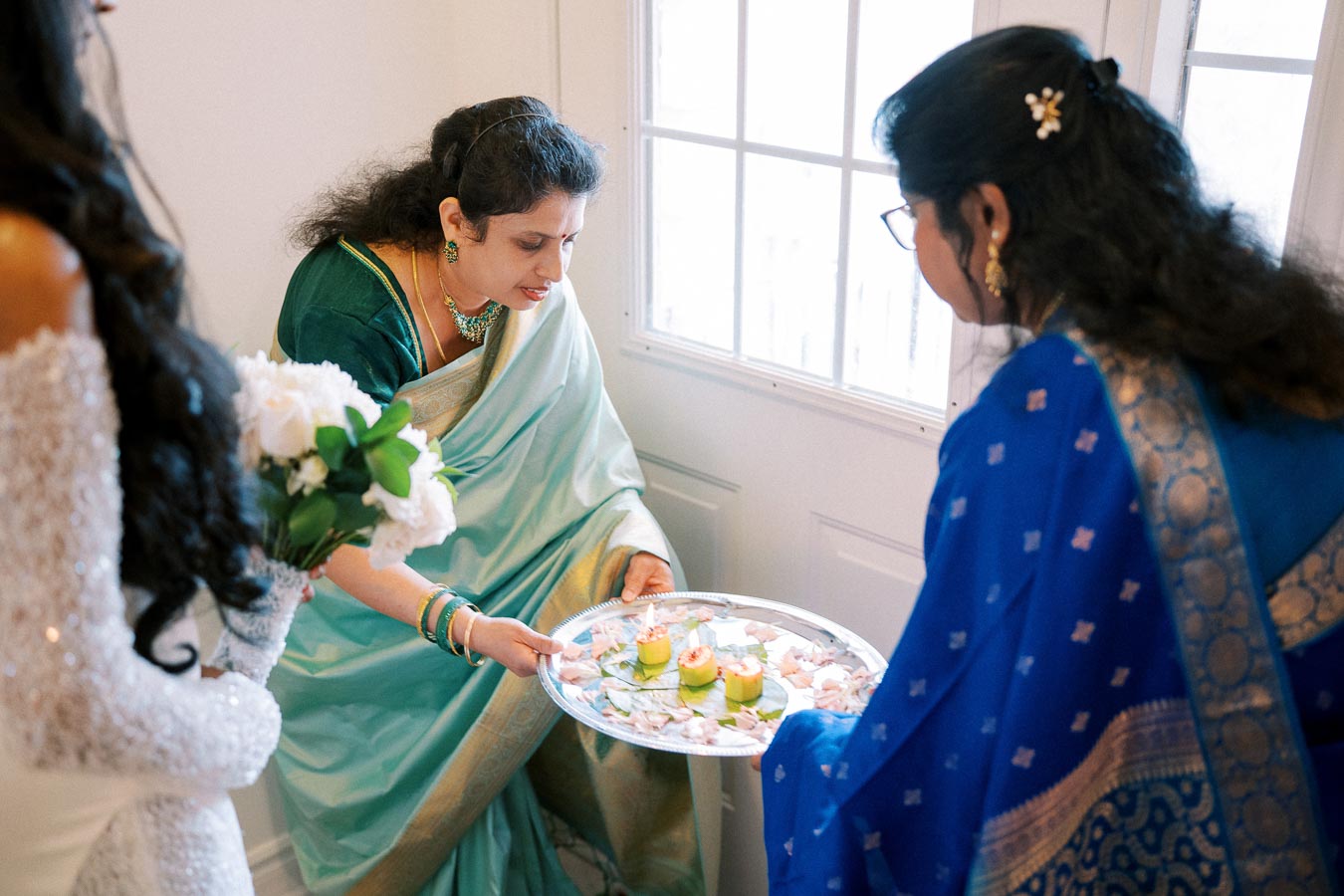 A woman in a green sari performs a traditional Indian ceremony, holding a decorated tray with candles and petals, as another woman in a blue sari looks on.
