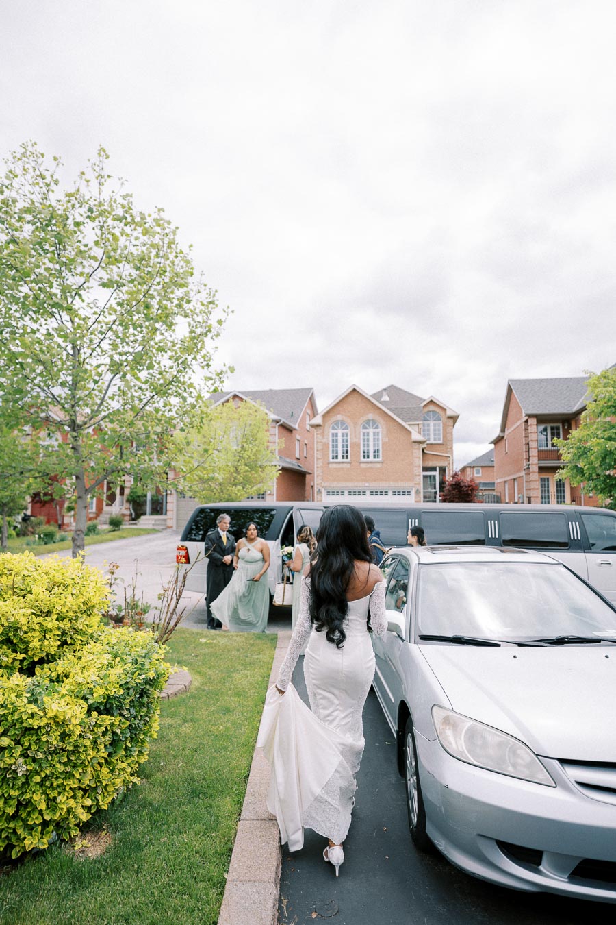 Bride in elegant white wedding dress walking towards a stretch limousine parked in a suburban neighborhood, surrounded by bridesmaids and lush green trees and bushes.