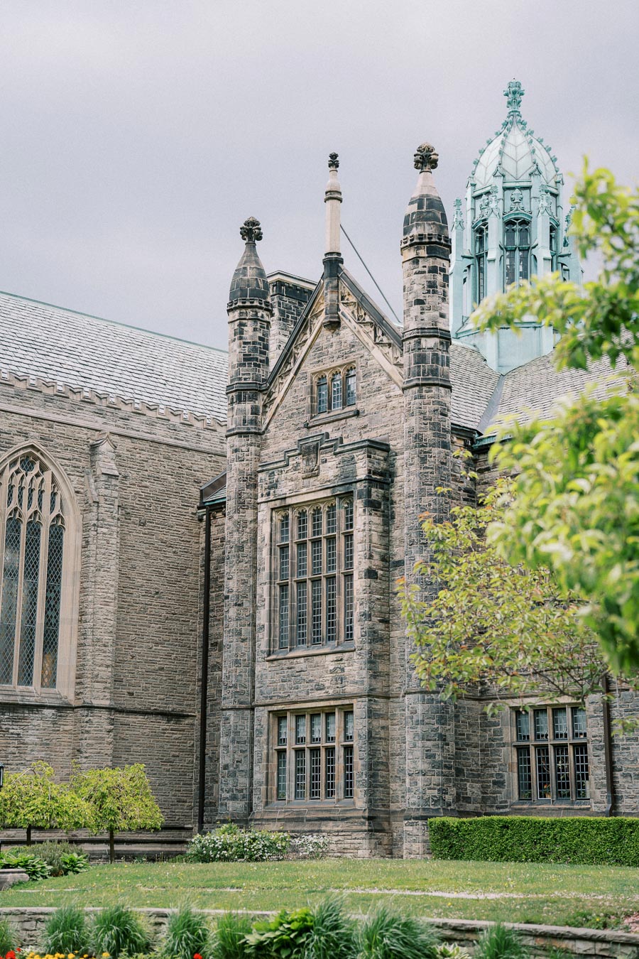 Gothic architectural style building with stone facade and tall windows, surrounded by lush greenery under an overcast sky.