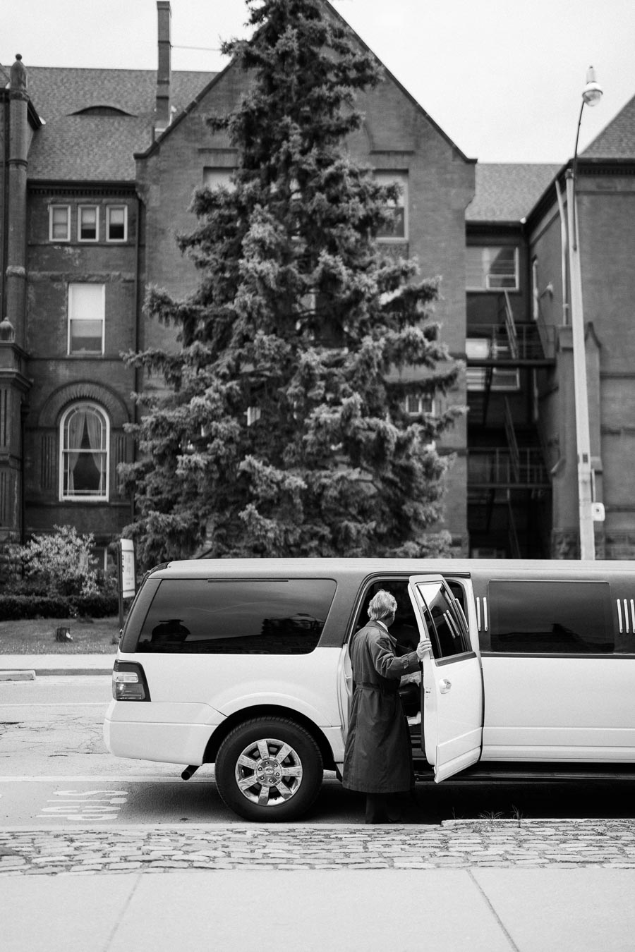 Black and white image of a person in a long coat entering a white stretch limousine parked in front of a historic brick building with large windows and a tall evergreen tree.