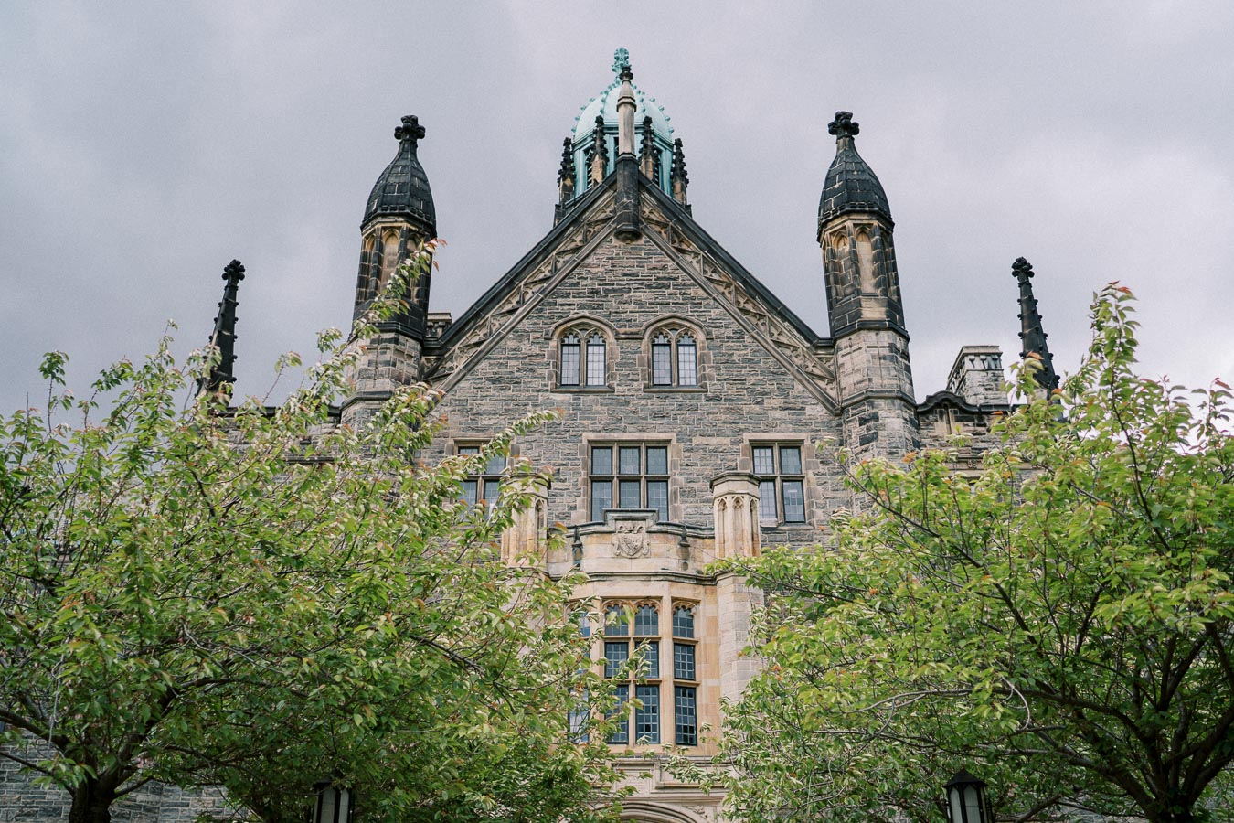 Historic Gothic Revival building with ornate architecture, featuring tall spires and intricate stonework, partially obscured by lush green trees under a cloudy sky.