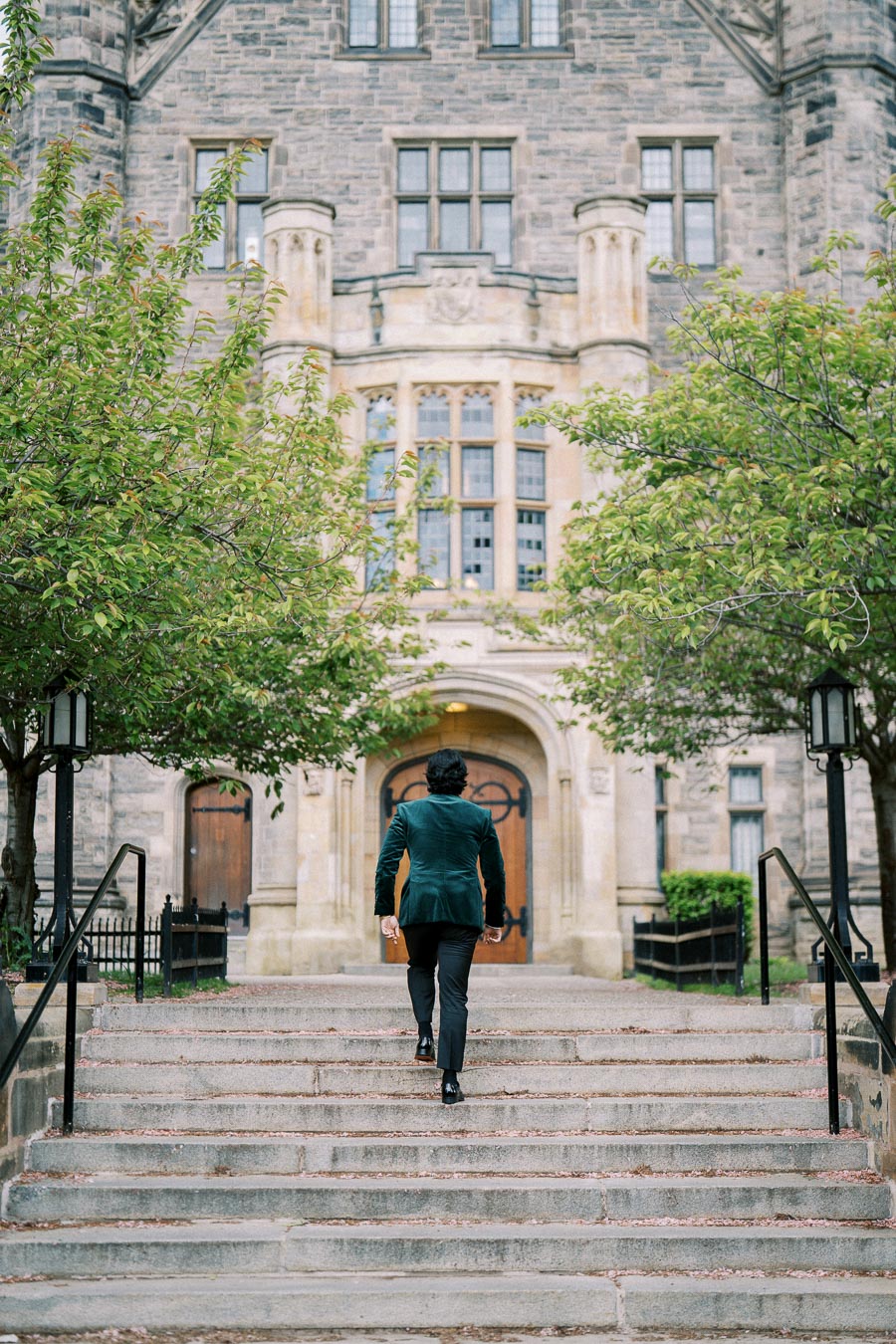 Man walking up stone steps towards historic building with arched wooden doors, surrounded by green trees, highlighting architectural design.