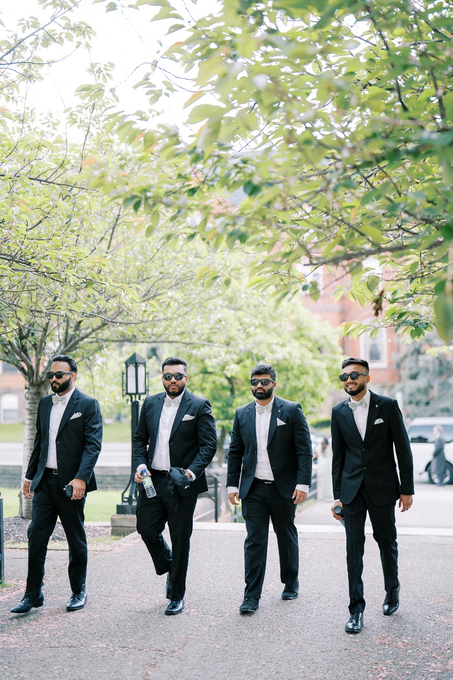 A group of four groomsmen in black suits and sunglasses walking through a scenic, tree-lined path on a sunny day, exuding a stylish and confident appearance.