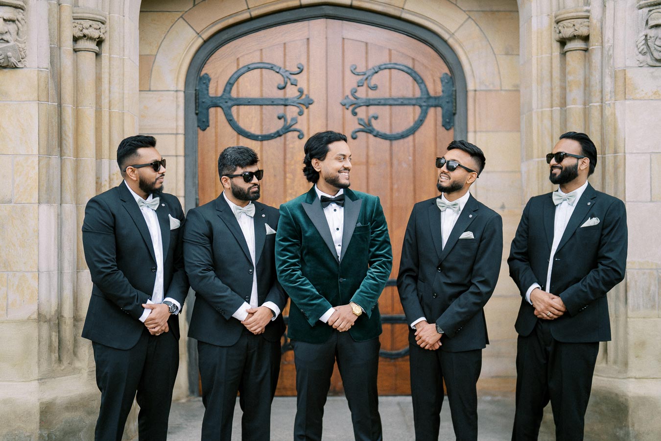 Group of five groomsmen in stylish suits and sunglasses standing together in front of a wooden arched door, smiling and posing at a wedding event.