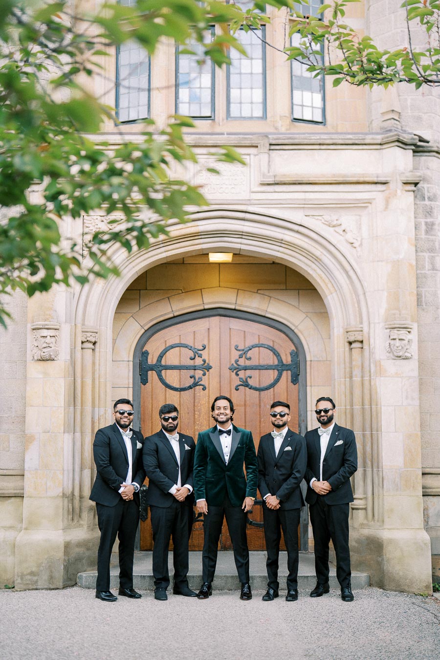 Group of five groomsmen in black suits and sunglasses posing in front of ornate wooden doors at a historic stone building, with greenery framing the scene.