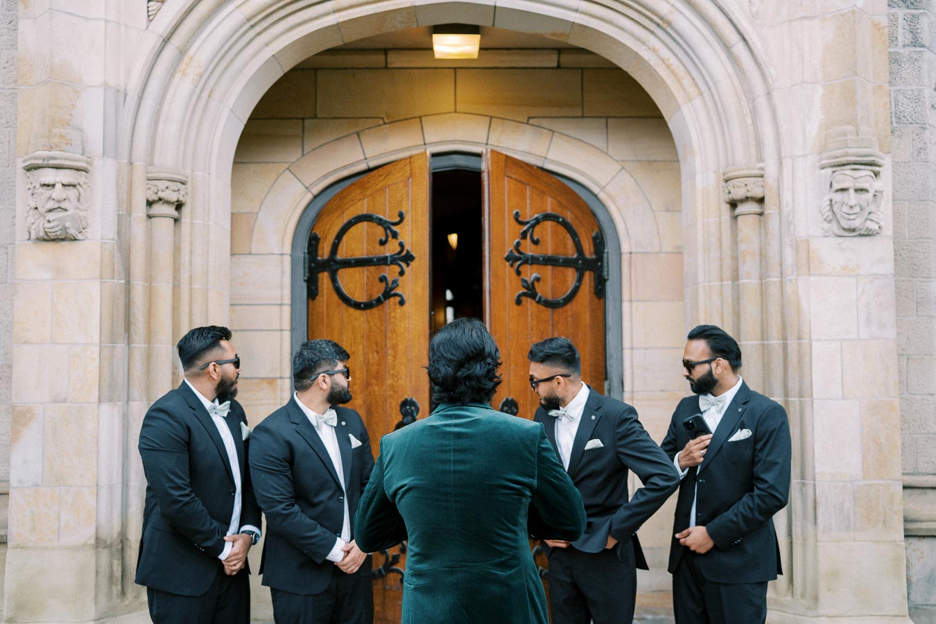 Groom and groomsmen in formal attire gather outside a stone church with ornate wooden doors before a wedding ceremony.
