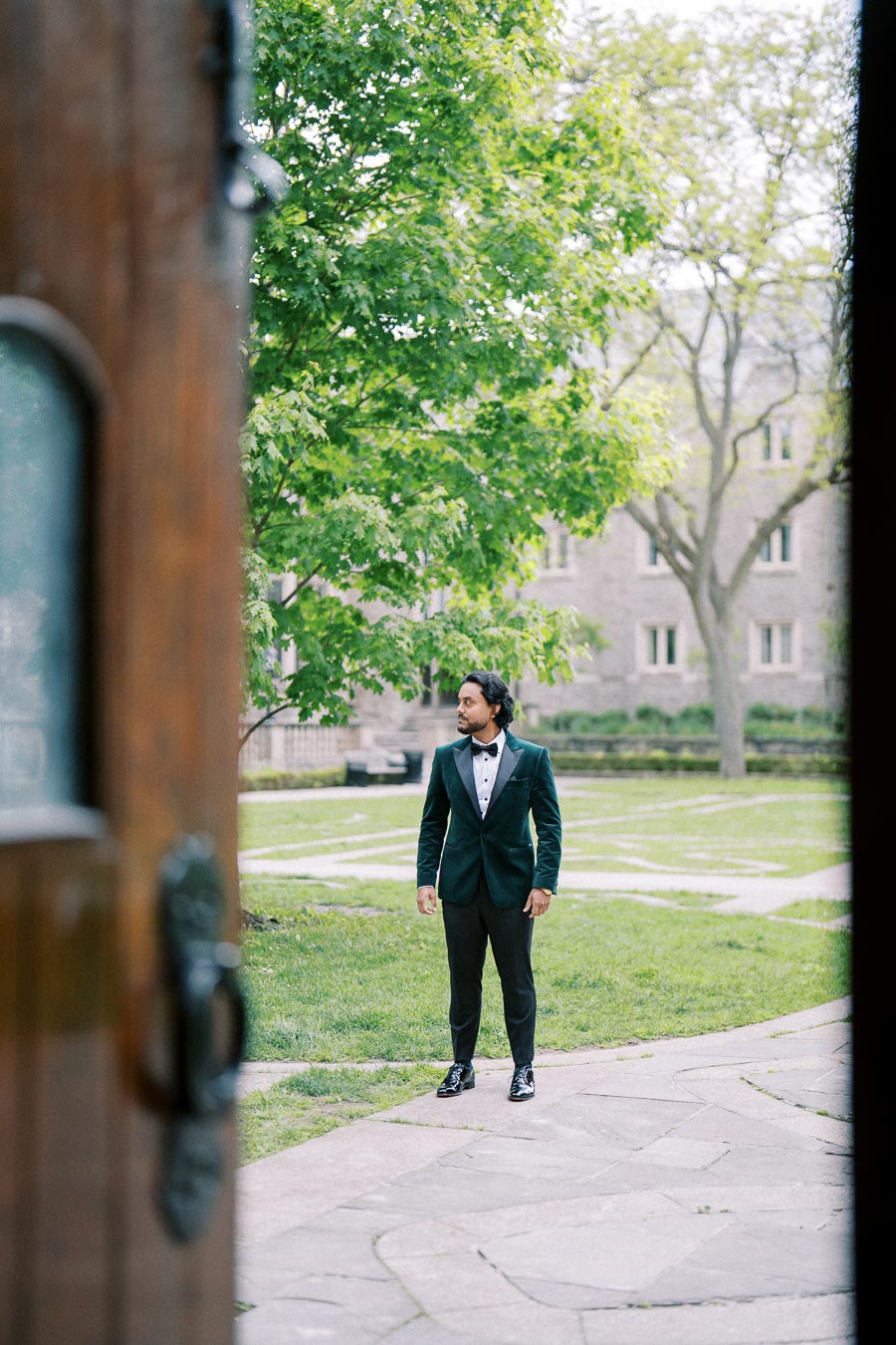 Man in a tuxedo standing outdoors on a stone pathway, surrounded by greenery, with a building in the background and a partially open door in the foreground.