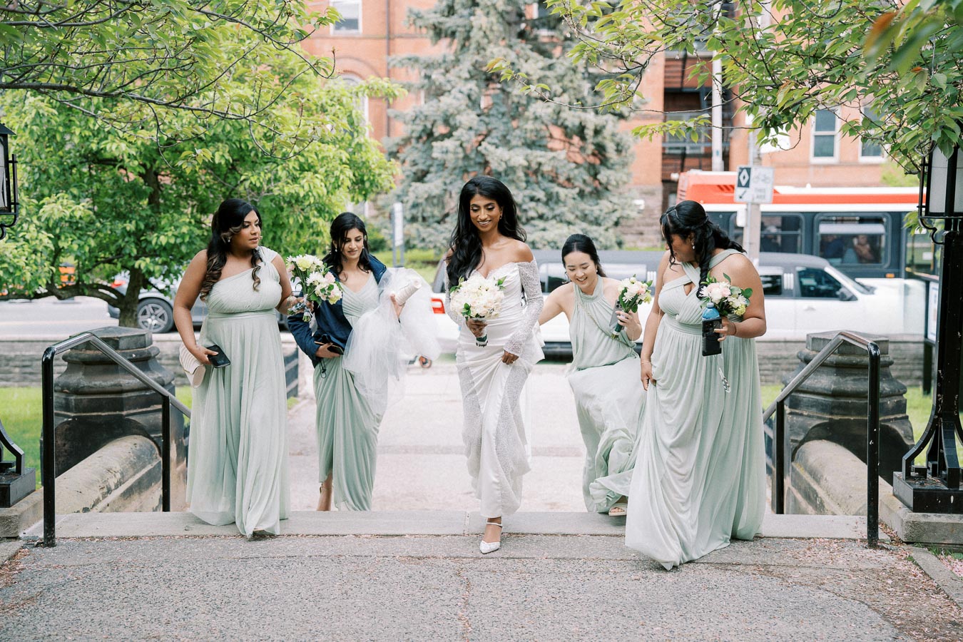 Bride accompanied by bridesmaids in light green dresses walking up outdoor steps, holding bouquets, with greenery and a street in the background.