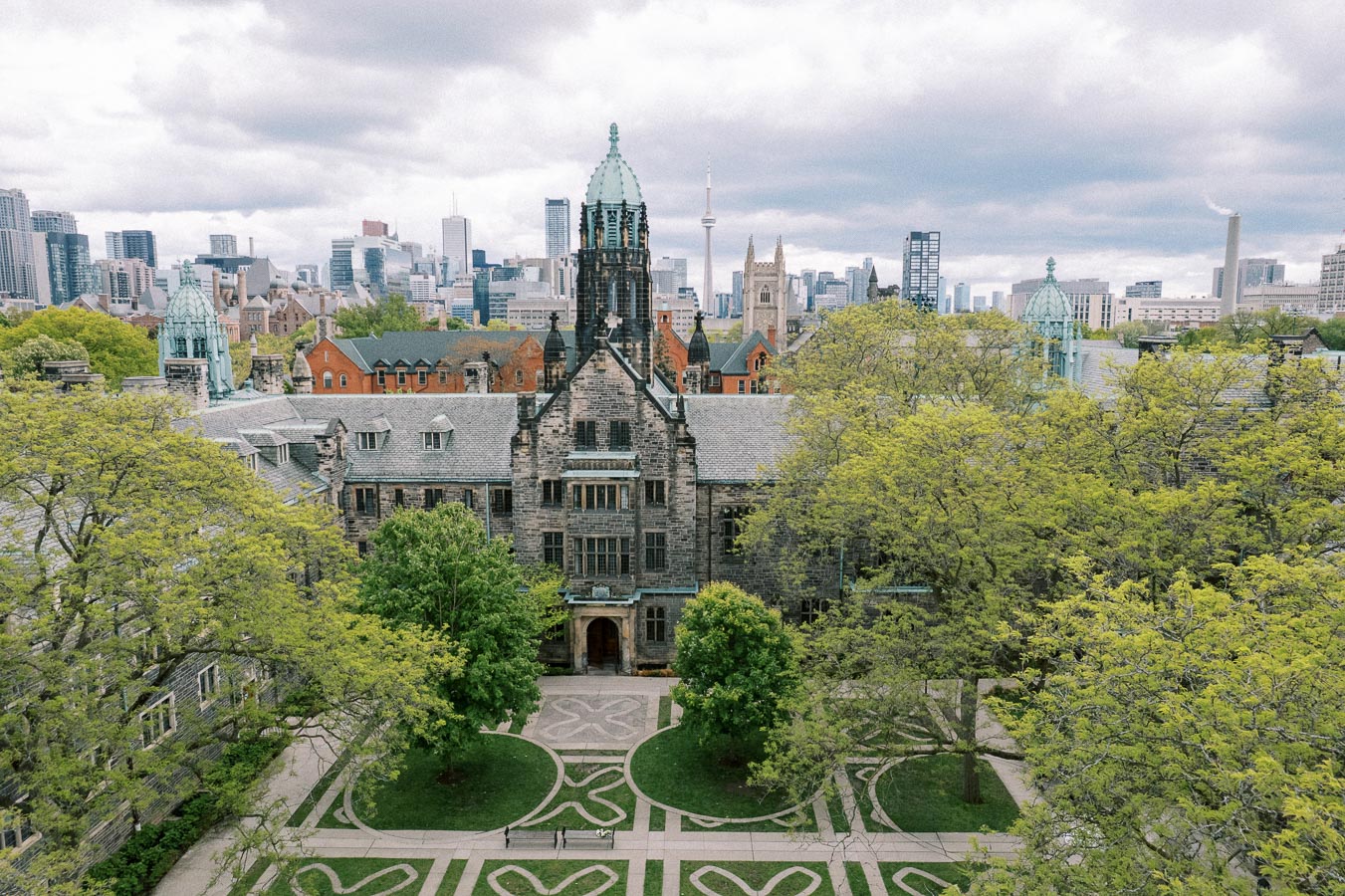 Aerial view of a historic university campus with a grand stone building surrounded by lush green trees, set against a city skyline under a cloudy sky.