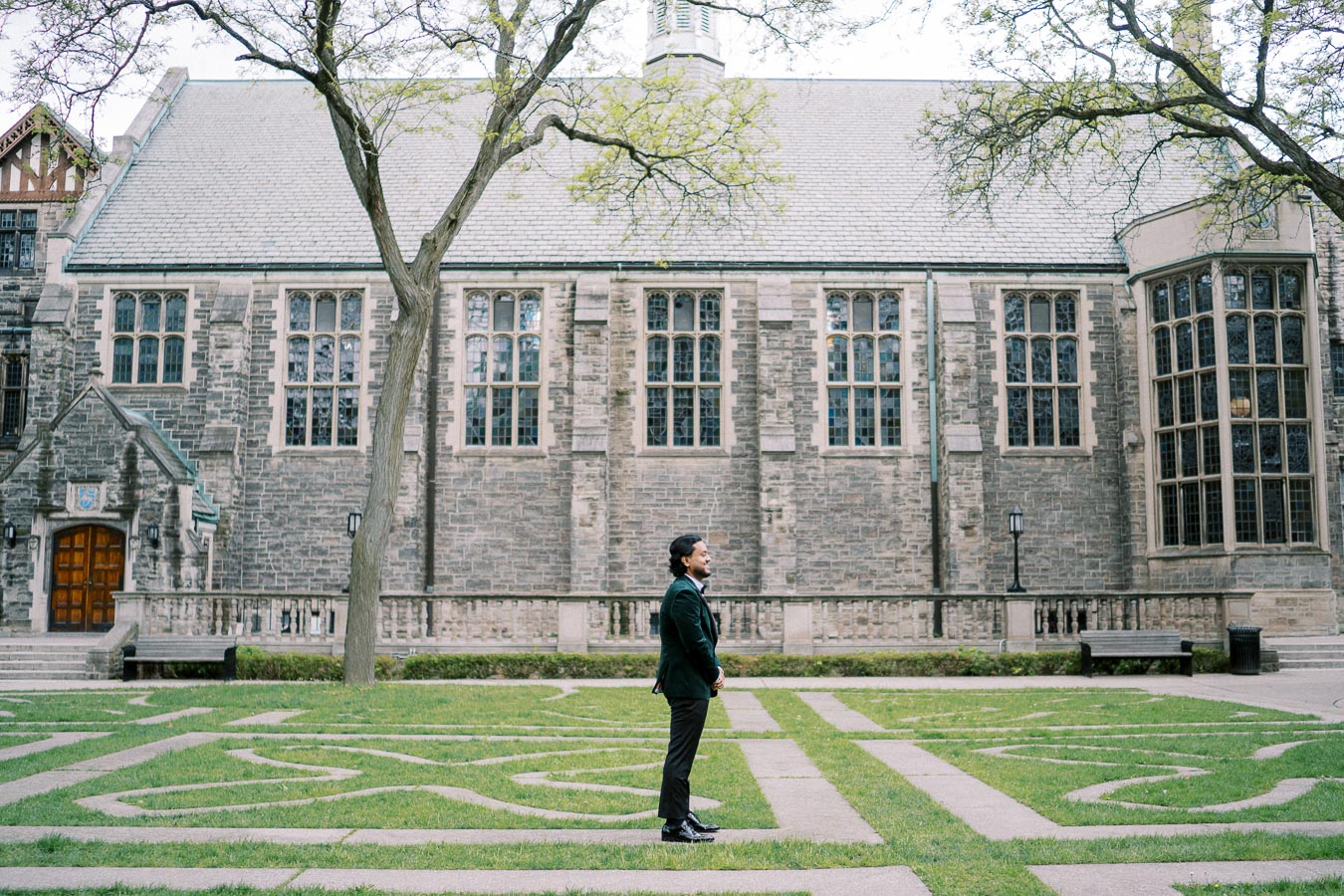 A person stands on a patterned lawn in front of a historic stone building with large windows and intricate architectural details, surrounded by trees.