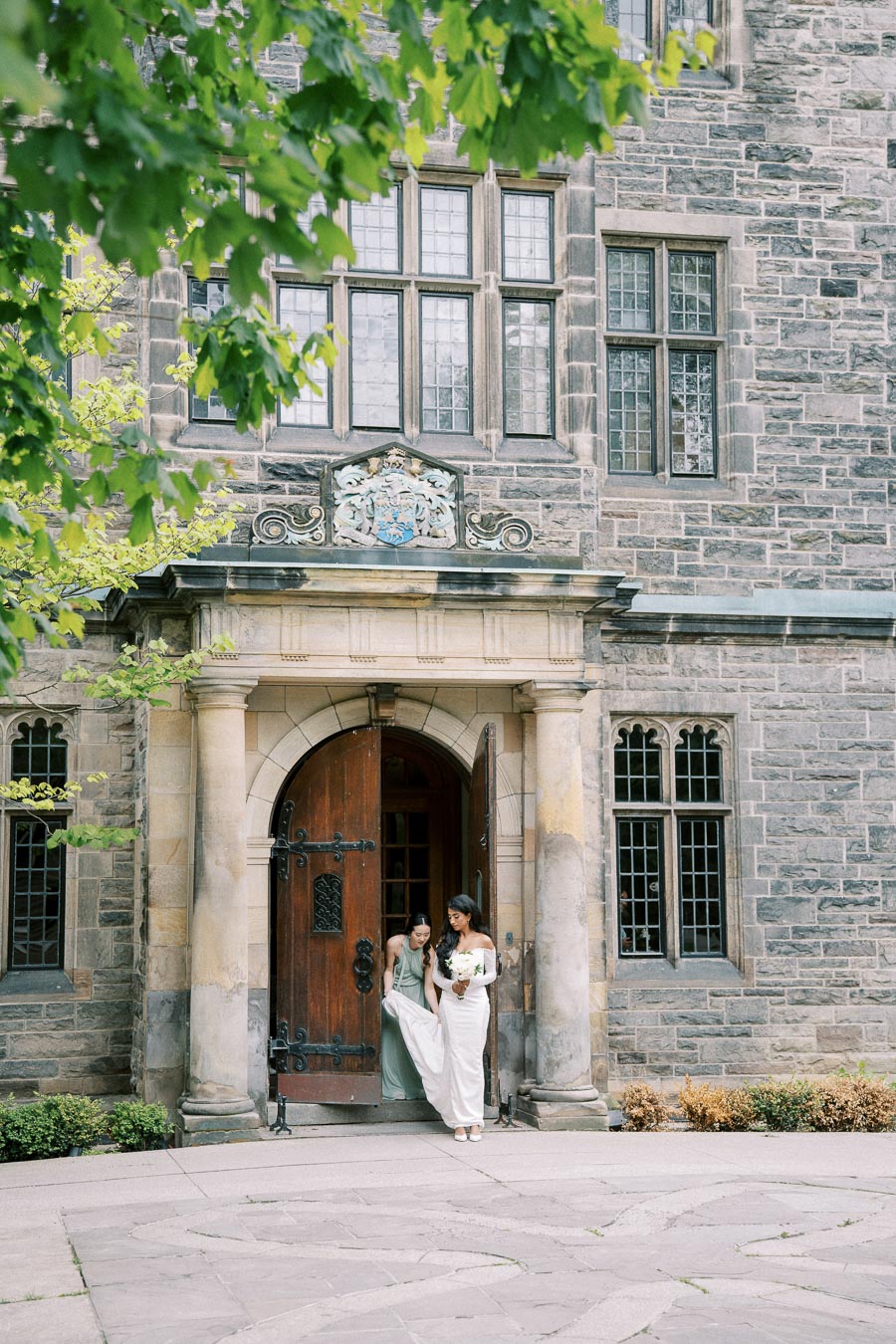 Two women in elegant dresses exit a historic stone building with a wooden arched door, surrounded by lush greenery, capturing a moment of celebration.