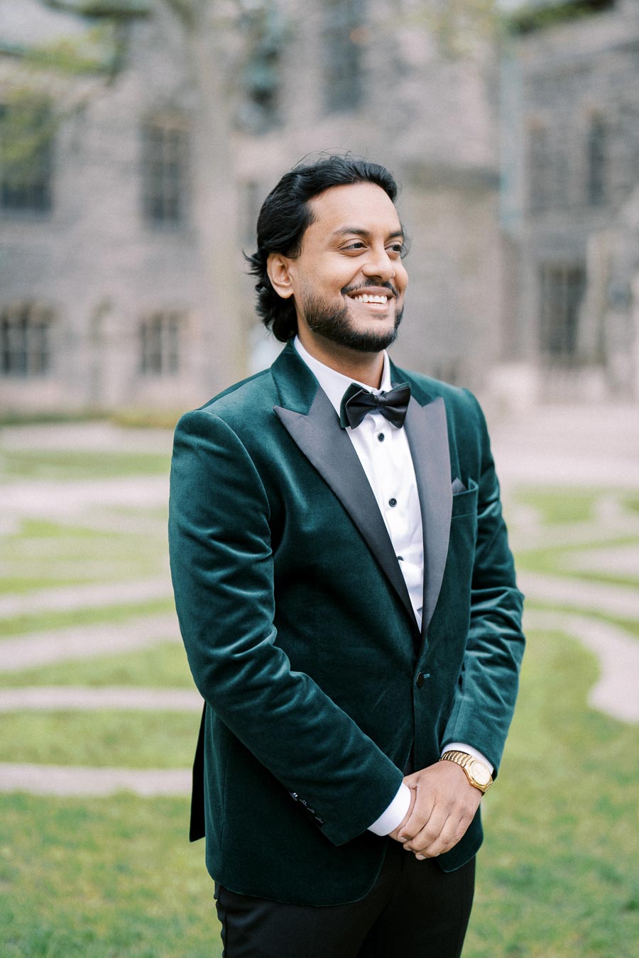 A man smiling in a formal green velvet suit and black bow tie, standing outdoors with a blurred background of building and grass.