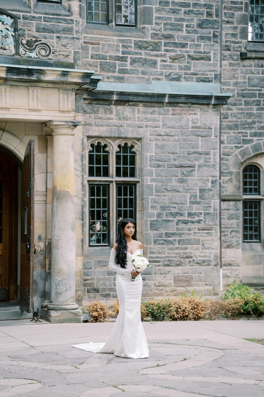 Bride in elegant white wedding dress holding bouquet stands in front of historic stone building, showcasing wedding fashion and architecture.