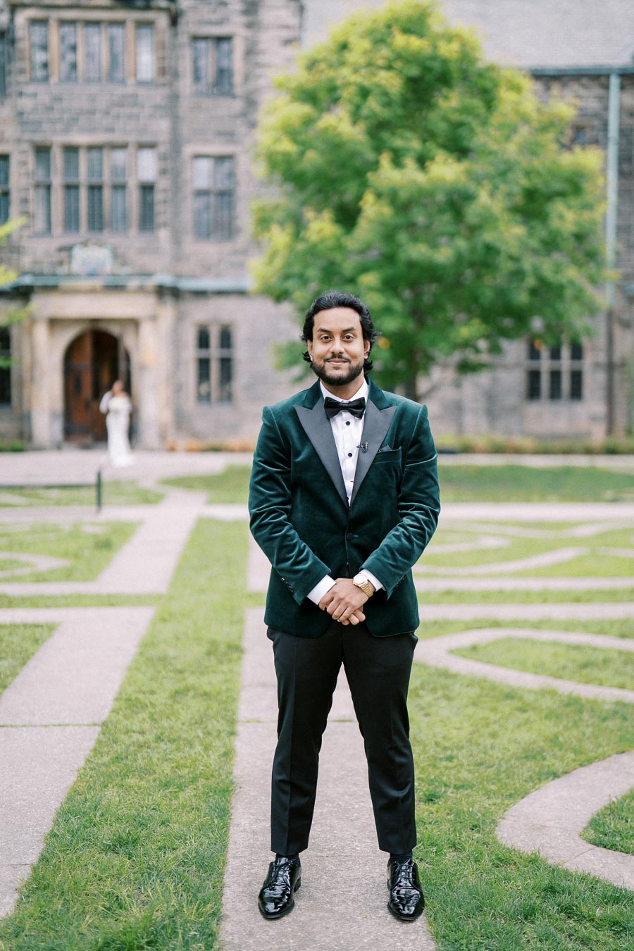 A man in a green velvet suit and bow tie stands confidently on a garden path in front of a historic stone building, with lush greenery and a tree in the background.