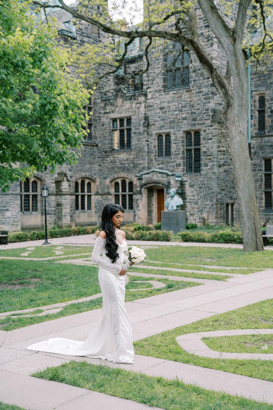 Bride in elegant white wedding dress holding bouquet, walking in a picturesque garden with historic stone building in the background.
