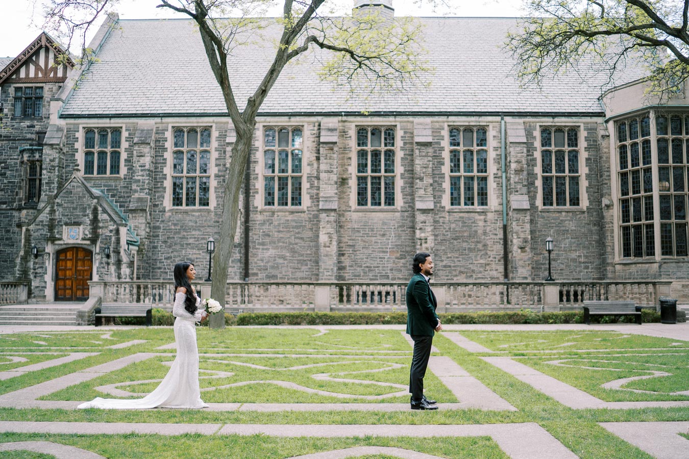 A bride in a white dress holds a bouquet, approaching a groom standing in a courtyard with a historic stone building in the background.