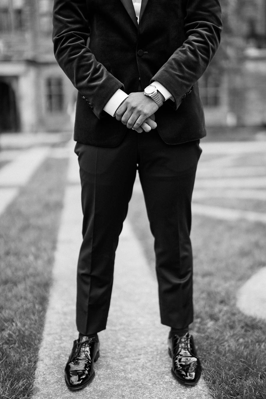 Black and white photo of a man in a suit, standing with hands clasped, showcasing formal attire and polished black shoes on a stone path.
