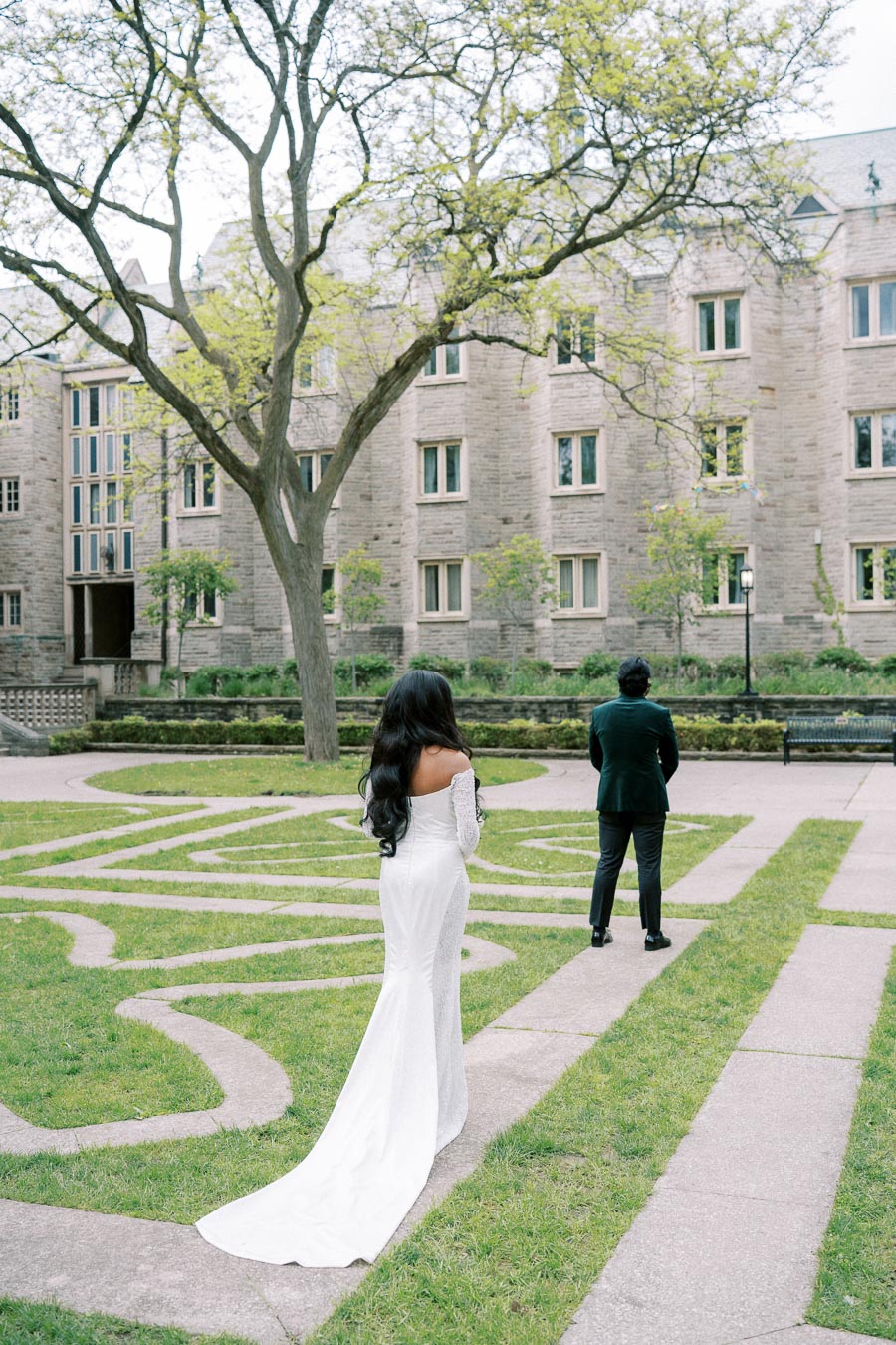 Bride in elegant white dress approaching groom for first look in lush green courtyard outside historic stone building.
