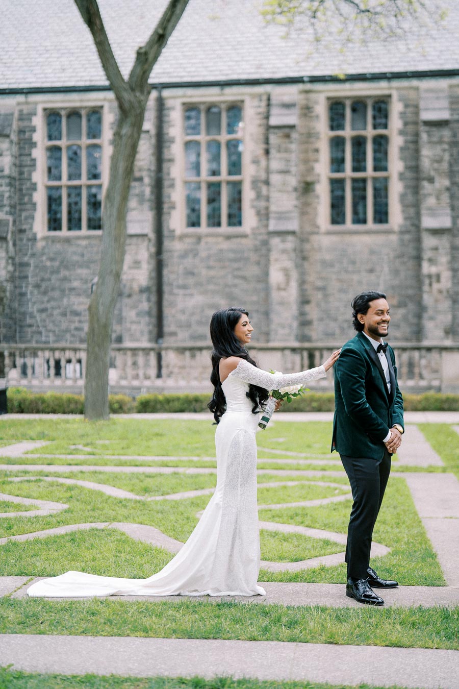 Bride in elegant white gown surprises groom in a tuxedo during their first look in a lush garden setting, with a historic stone building in the background.