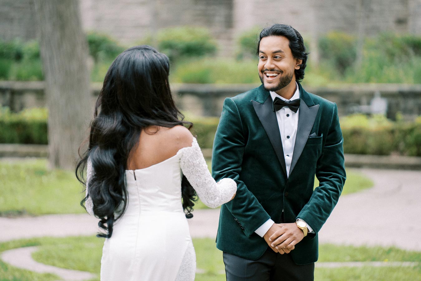 A bride in a white dress and a groom in a green suit share a joyful moment in a garden setting.