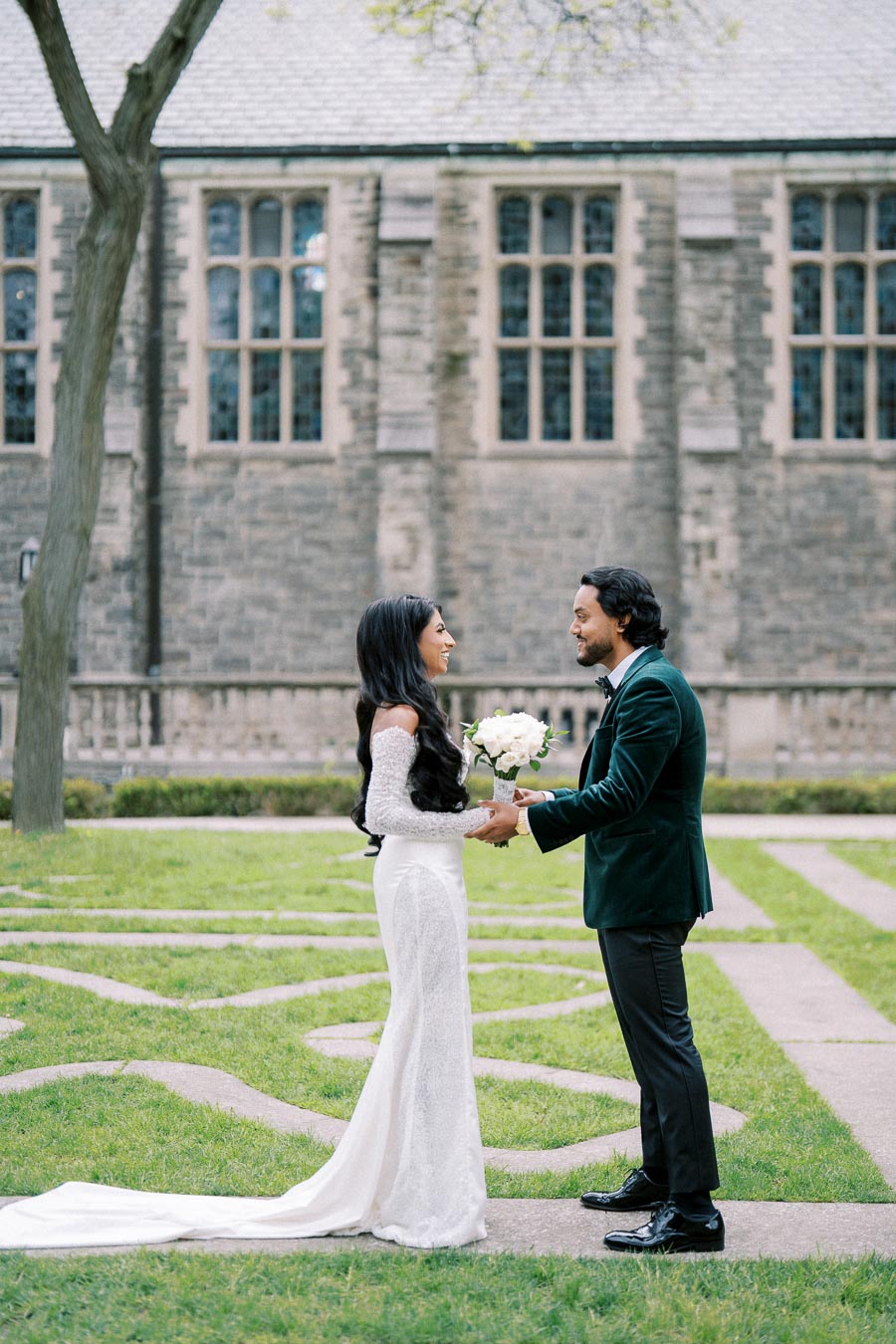 A couple stands facing each other outdoors in front of a historic stone building. The woman is in a long, elegant white dress holding a bouquet of flowers, and the man is wearing a dark suit. The lush green grass and trees enhance the romantic atmosphere of the scene.