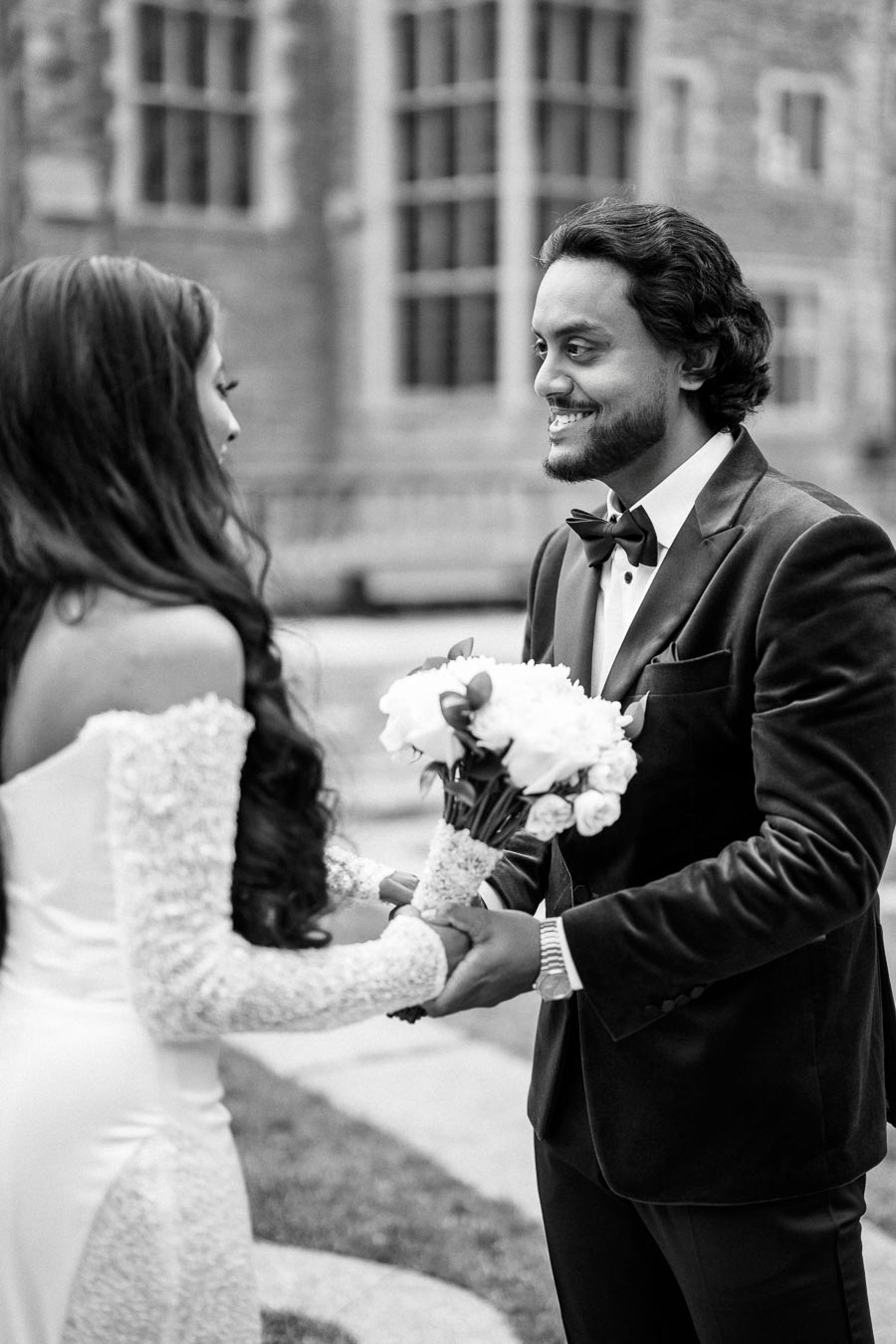 Black and white photo of a bride and groom holding hands and smiling at each other outdoors, with the bride holding a bouquet of flowers, the groom wearing a tuxedo, and an elegant building in the background.