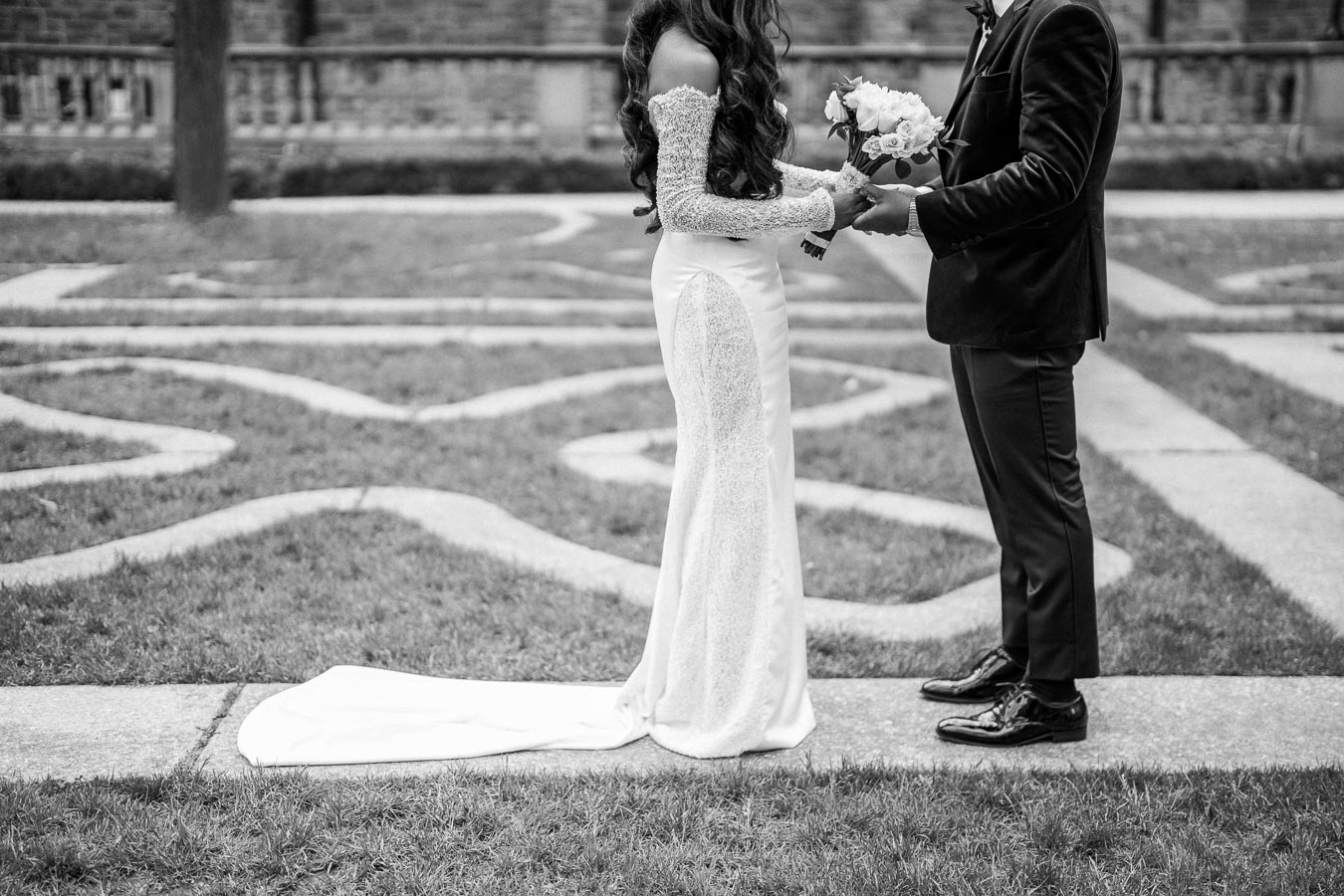 A black and white image of a bride and groom holding hands with a bouquet, standing in an outdoor garden setting, showcasing a beautiful wedding dress with a long train.