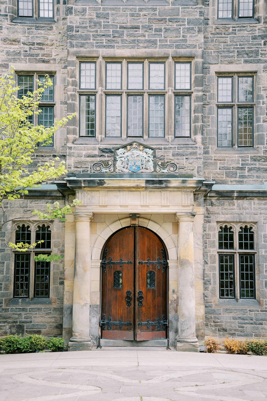 Historic building entrance with a large wooden door and intricate stonework, featuring medieval windows and ornate architectural details. Ivy partially covers the surrounding stone facade, adding a classic charm.