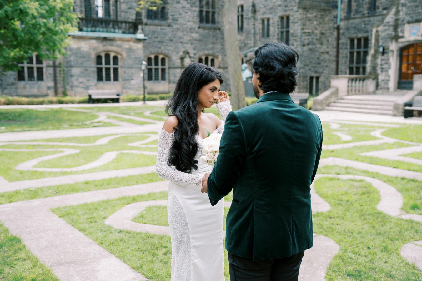 A bride in a white gown and a groom in a green suit share a moment in a picturesque garden setting with a historic stone building in the background.