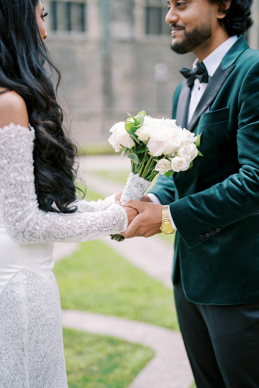 A bride and groom holding hands with a bouquet of white roses, standing outdoors in a garden setting. The bride is wearing an elegant off-the-shoulder wedding dress, and the groom is in a formal dark green velvet suit.