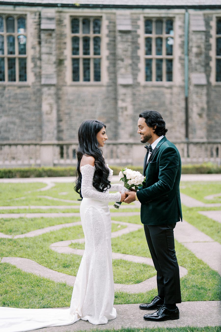 A bride and groom holding hands and exchanging vows in an outdoor courtyard setting, with the bride in a white lace gown and the groom in a dark green velvet suit, set against a stone building backdrop.