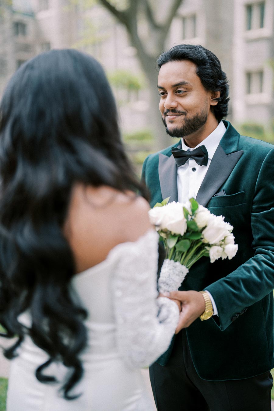 A groom in a green velvet tuxedo holds a bouquet of white roses, smiling at a bride in a white gown during their wedding ceremony.