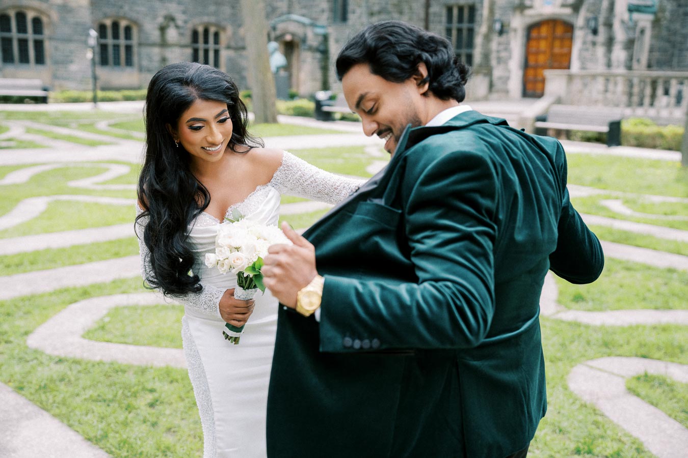 A bride in a white wedding dress holding a bouquet smiles at a groom in a green velvet suit at an outdoor venue with stone pathways and historic architecture in the background.