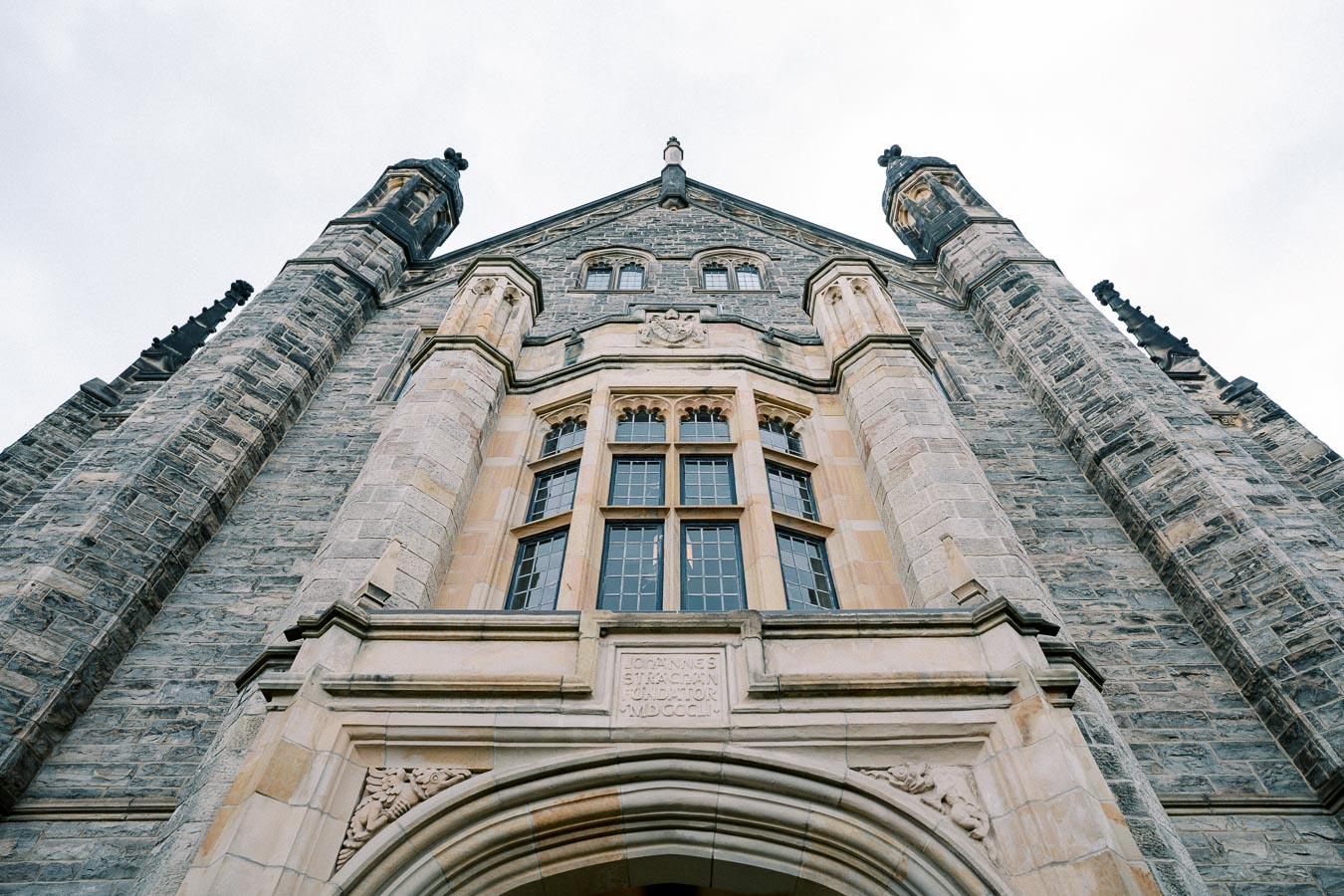 Upward view of a historic stone building with gothic architecture, featuring ornate windows and detailed stonework against a cloudy sky background.