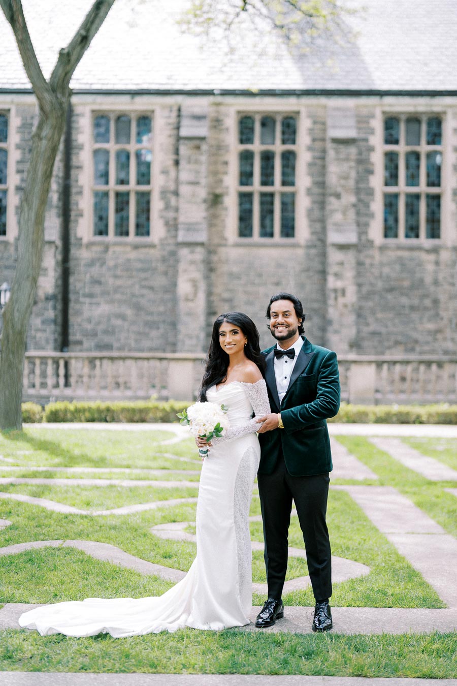 Bride and groom posing outdoors in elegant wedding attire, with a historic stone building in the background, surrounded by lush green grass.