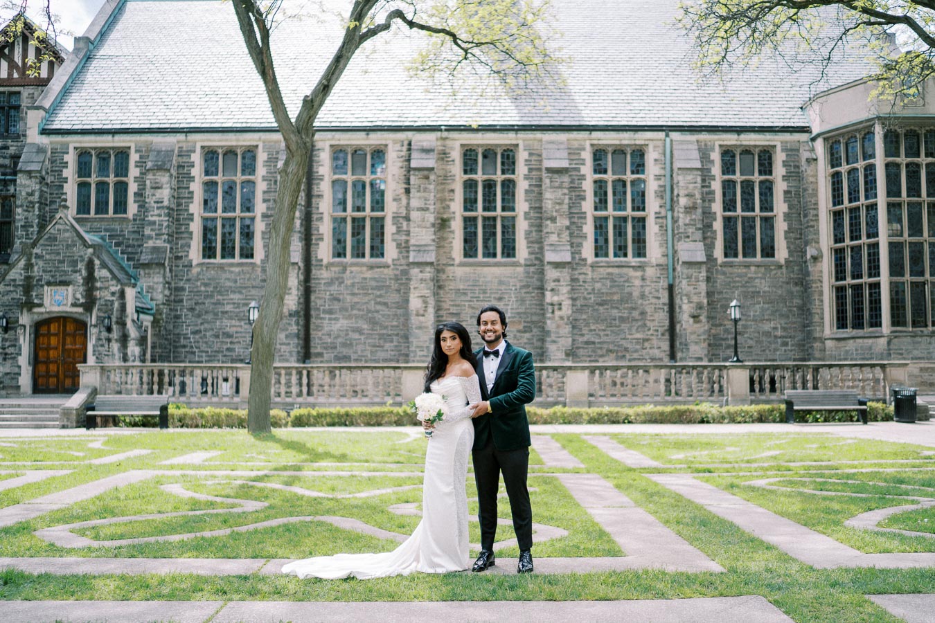 Elegant bride and groom posing in front of a historic stone building with arched windows, set in a beautifully manicured garden, perfect for a wedding photo shoot.