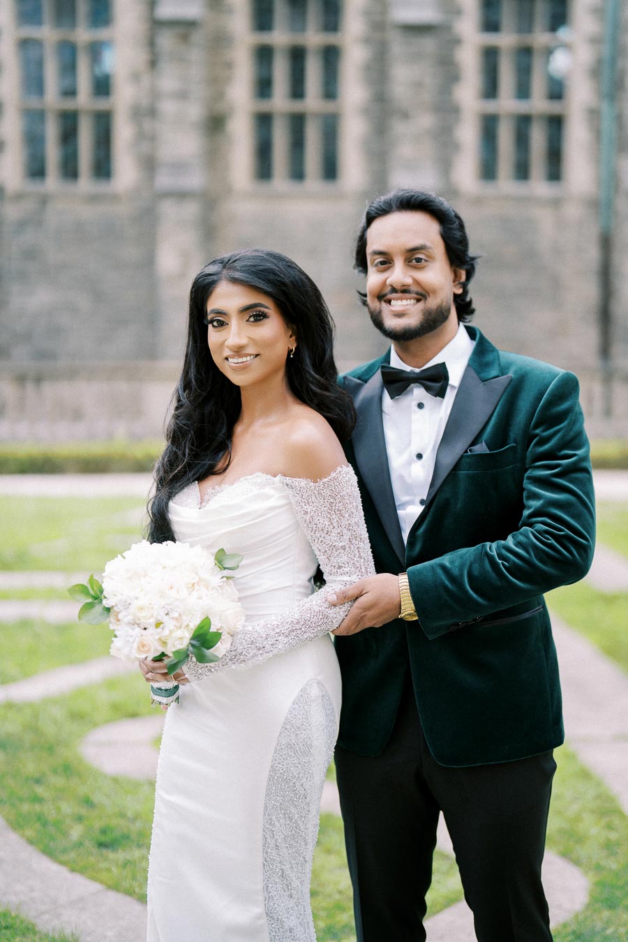 Elegant bride and groom posing outdoors, bride holding a bouquet of white flowers in a scenic garden setting with historic stone architecture in the background.
