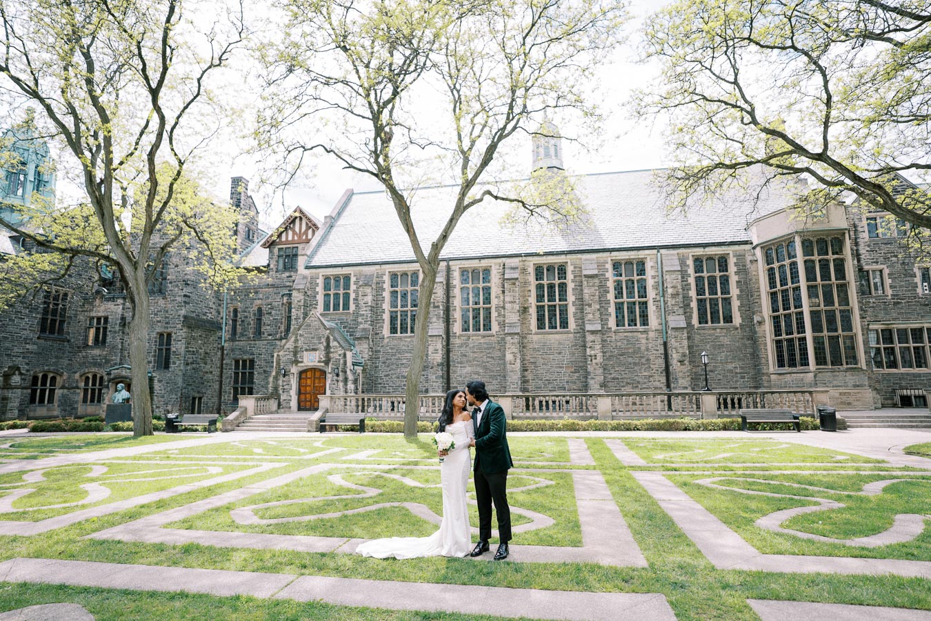 Bride and groom embracing in historic courtyard with ornate stone building and lush green lawn, featuring intricate path design and tall trees.
