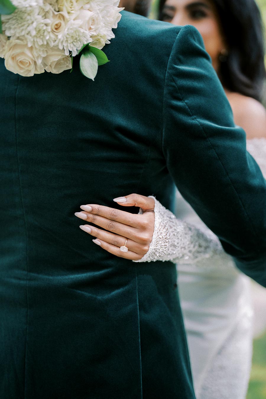 A close-up of a bride and groom embracing, showcasing the bride's hand with a diamond ring resting on the groom's dark green velvet suit. A bouquet of white flowers complements the elegant scene.