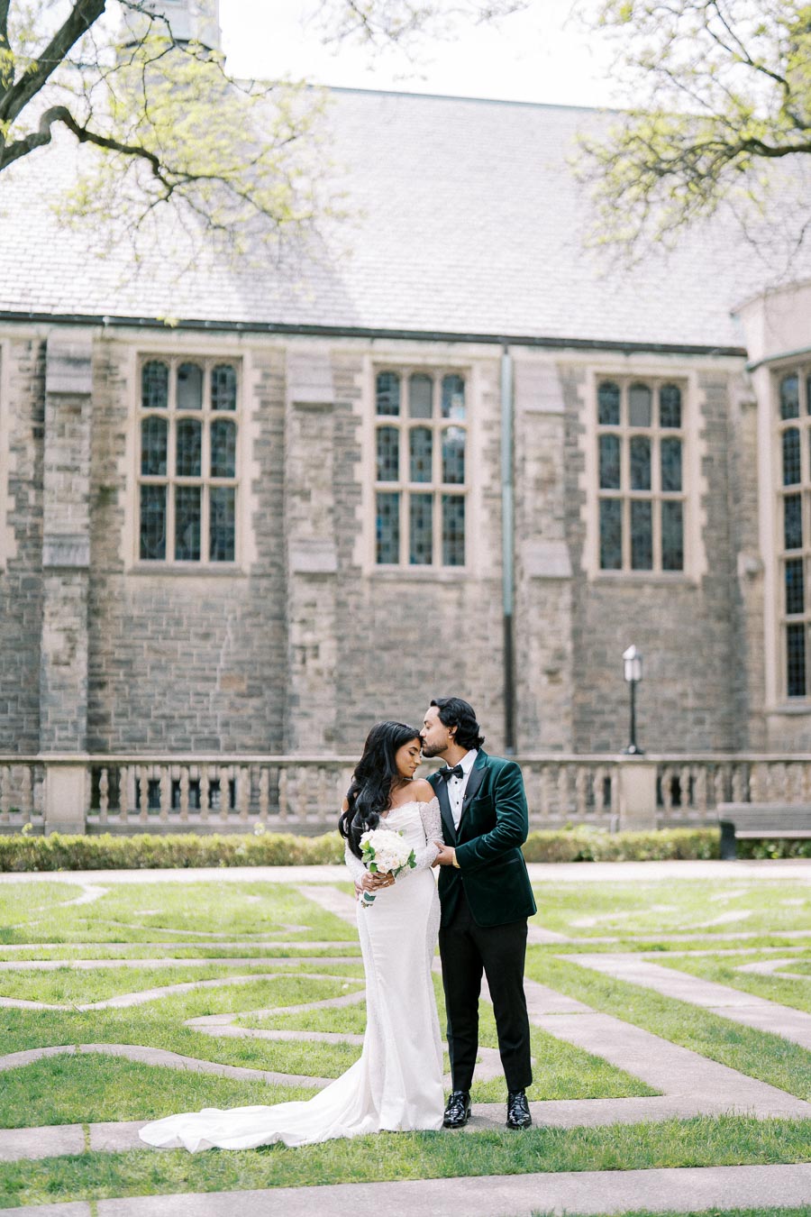 A bride and groom share a romantic moment during their wedding photoshoot in front of a historic stone building, surrounded by greenery. The bride, in an elegant off-the-shoulder white gown, holds a bouquet of flowers while the groom, in a stylish dark green jacket and black trousers, lovingly kisses her forehead.