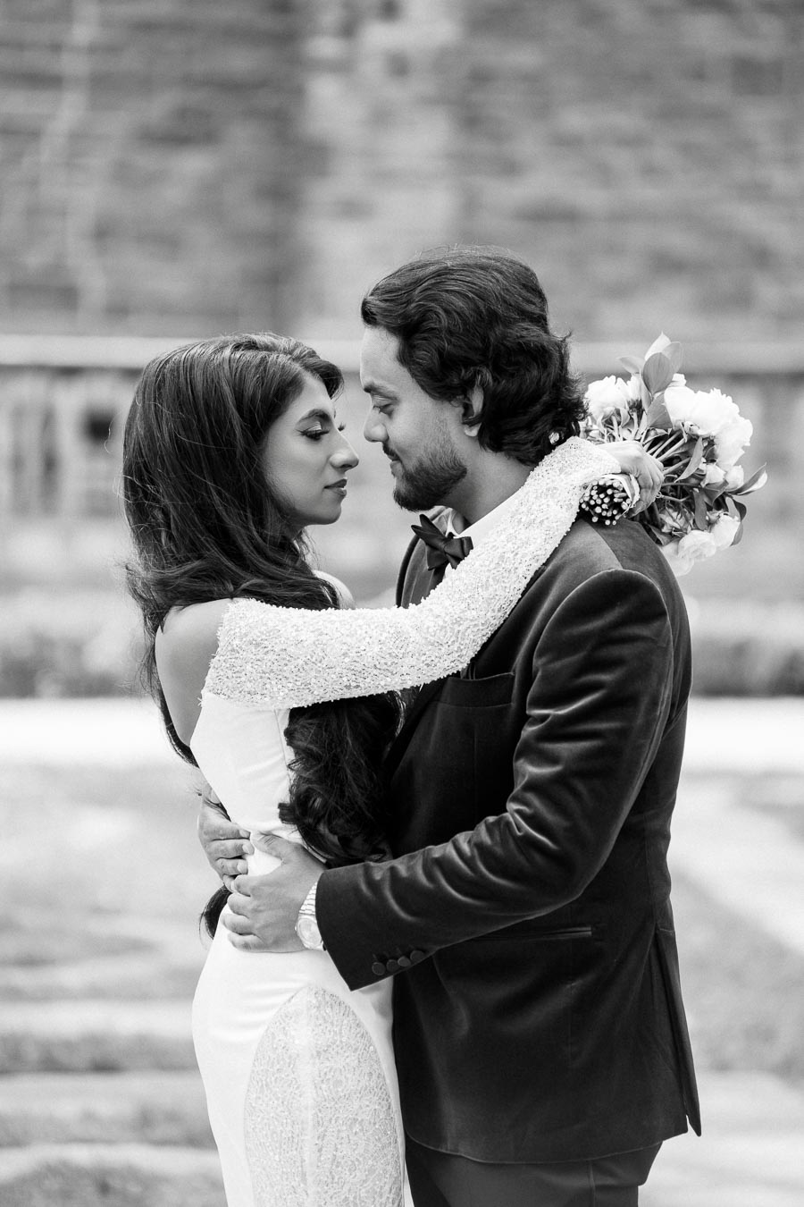 A romantic black and white photo of a couple embracing, with the woman holding a bouquet of flowers, set against a blurred outdoor background.