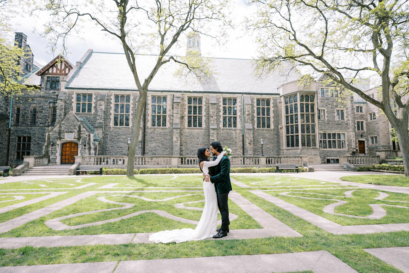 A romantic couple embraces in front of a historic stone building with tall windows, surrounded by manicured gardens and trees on a sunny day.