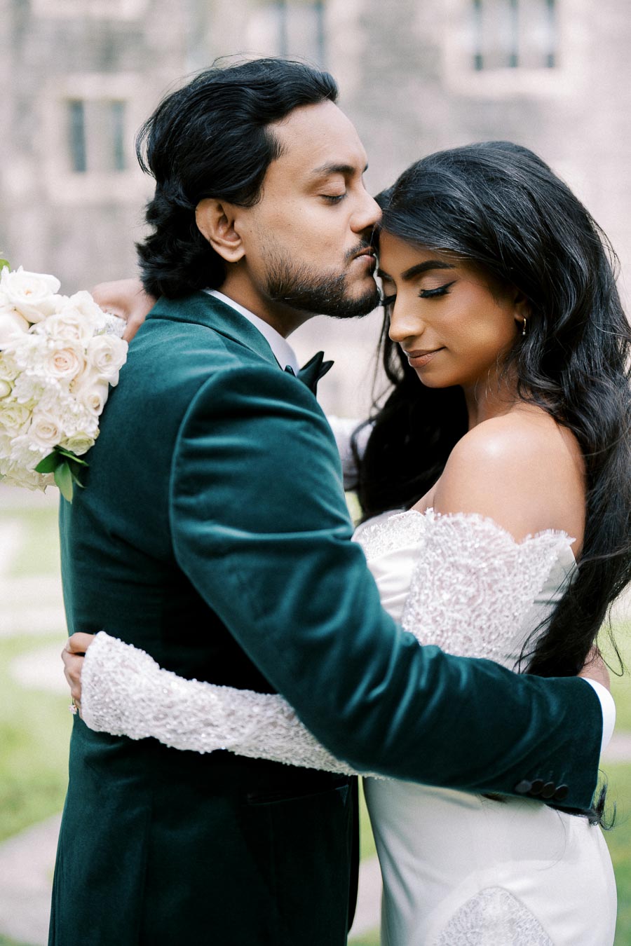 A bride and groom embrace tenderly in an outdoor setting, with the groom lovingly kissing the bride's forehead. The bride holds a bouquet of white roses, wearing a lace wedding dress, and the groom is in a dark teal suit.