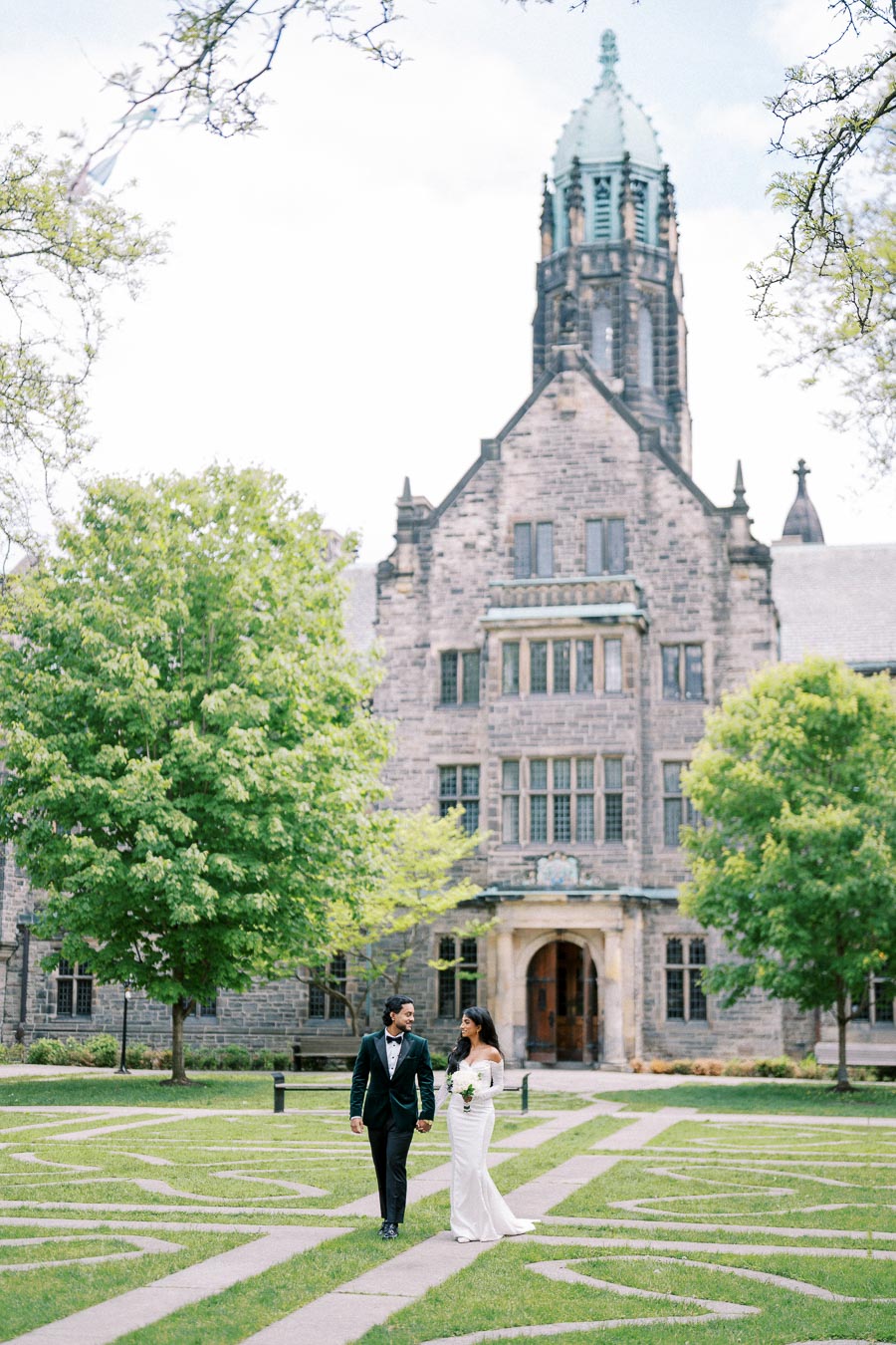 A bride and groom walking hand in hand on a green lawn in front of a historic stone building, surrounded by trees on a bright day.