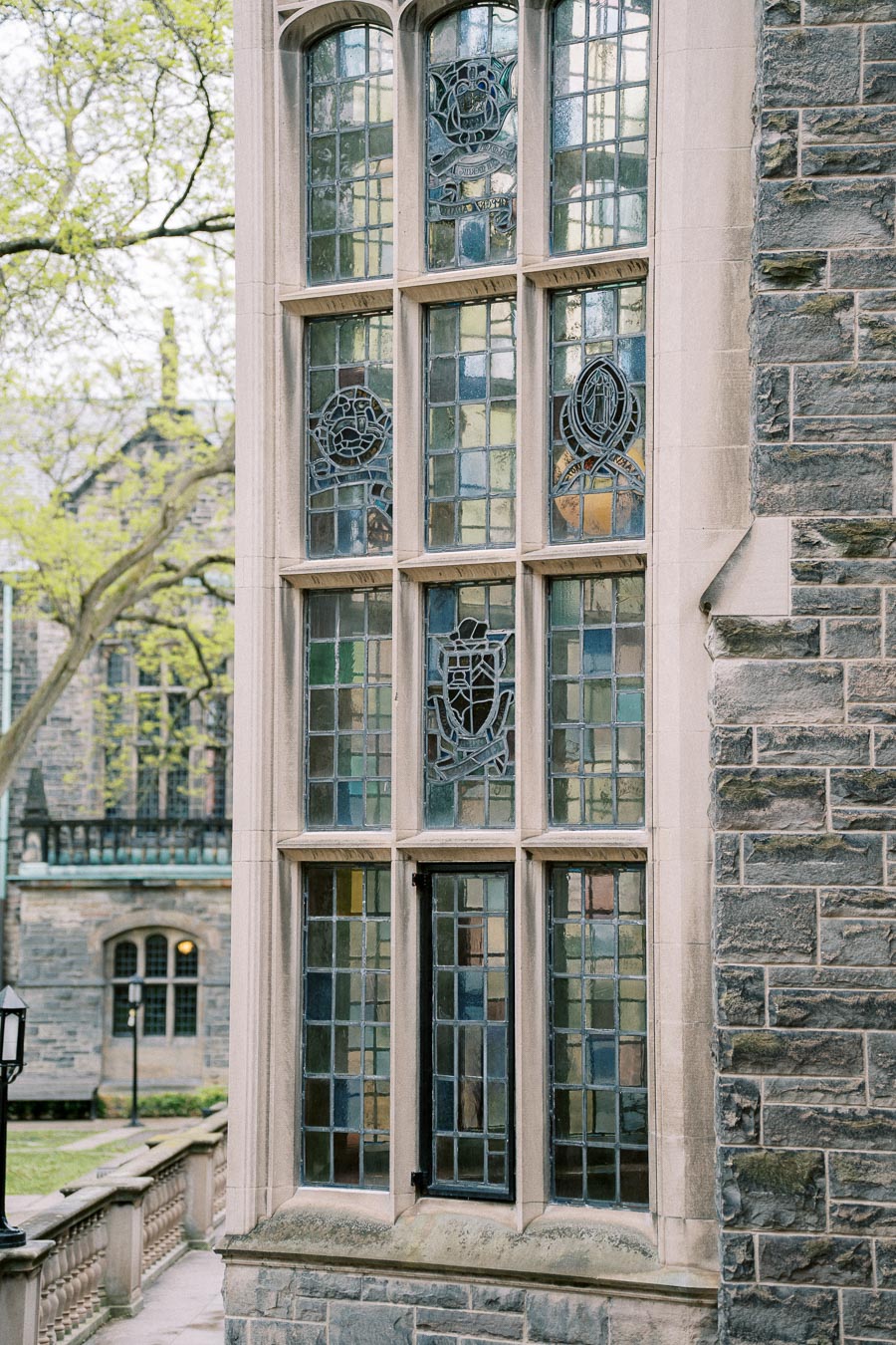 Stained glass window on a historic stone building facade, surrounded by lush green trees and an ornate railing.