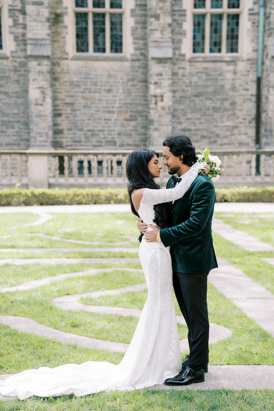 Elegant couple embracing in wedding attire with a stone building backdrop, bride in white lace gown and groom in dark suit.