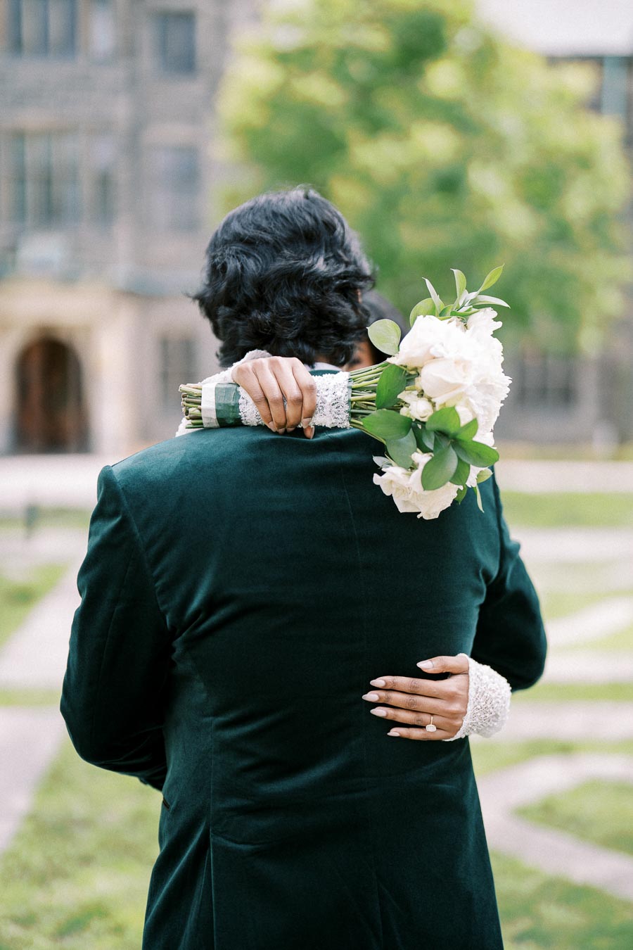 A bride embraces her partner from behind, holding a bouquet of white flowers wrapped in lace, with a visible engagement ring. The couple stands outdoors in front of a historic building with greenery in the background, creating a romantic wedding scene.