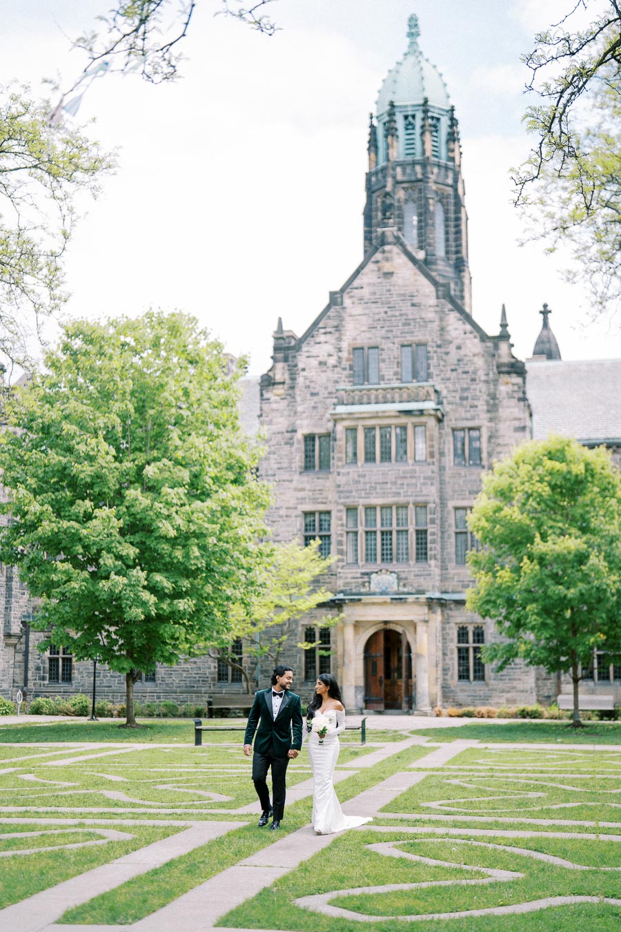 Wedding couple walking on picturesque stone pathway with historic stone building and lush green trees in the background, creating a romantic and elegant scene.