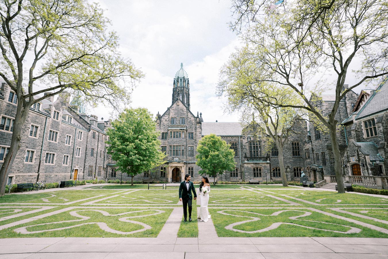 A couple elegantly dressed in wedding attire walks hand in hand across a beautifully landscaped courtyard with intricate grass patterns, surrounded by historic stone buildings and tall trees on a sunny day.